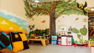 Une salle de jeux pour enfants avec une fresque représentant un arbre, des jouets et une cuisine miniature.