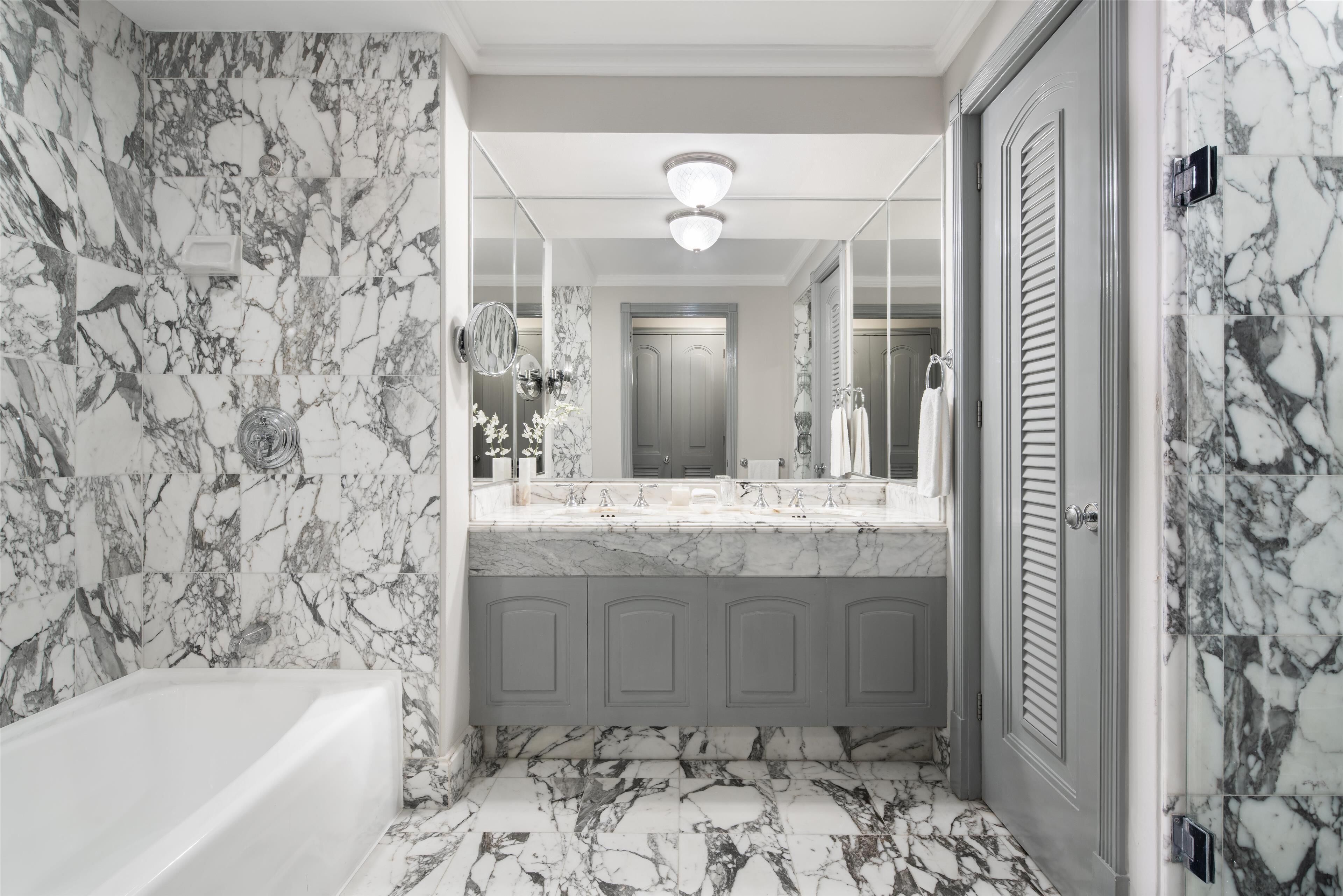 A bathroom with marble walls and floor, a bathtub, a vanity with a mirror, and a door with louvered panels.