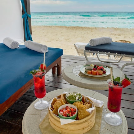 A beachside dining setup with a table set for two, featuring a fruit salad, chips, and a drink.