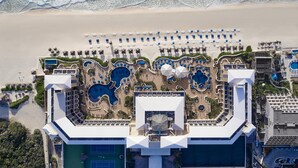 Vista aérea de un resort con varias piscinas, una zona de playa con sombrillas y un edificio grande con un techo distintivo.
