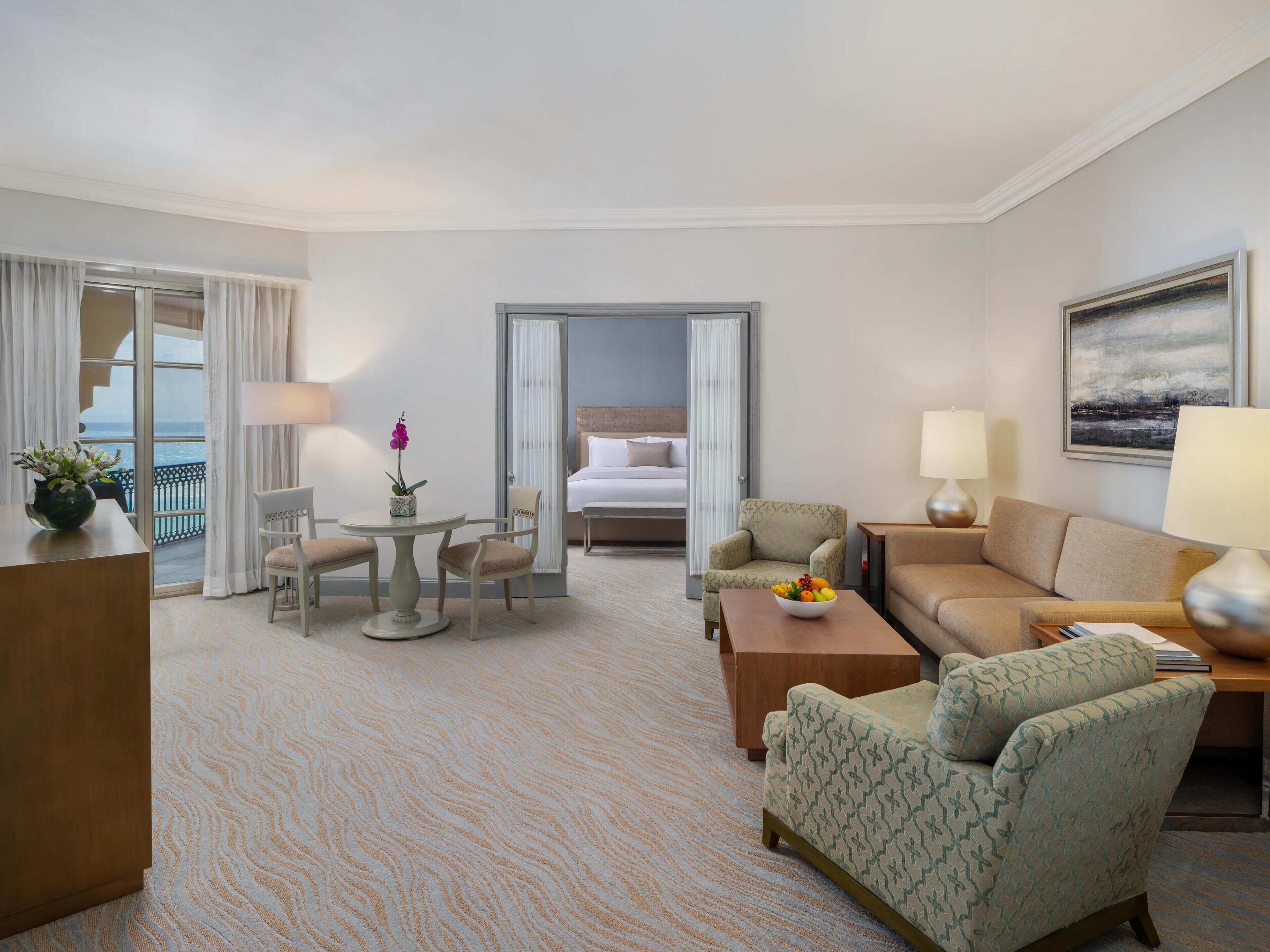 Quarto de hotel com cama, área de estar com mesa e cadeiras, sofá e vista para o oceano através de amplas janelas.