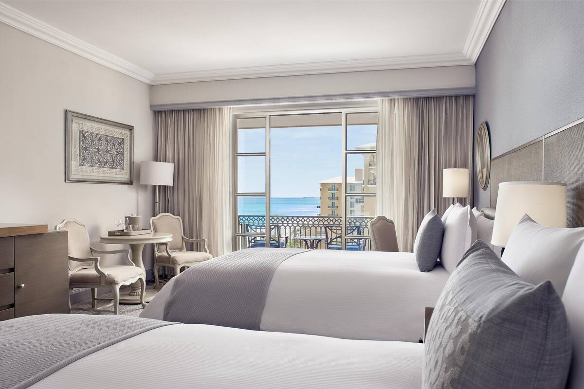 Une chambre d’hôtel avec deux lits, un bureau, une chaise, une table et un balcon donnant sur la mer.