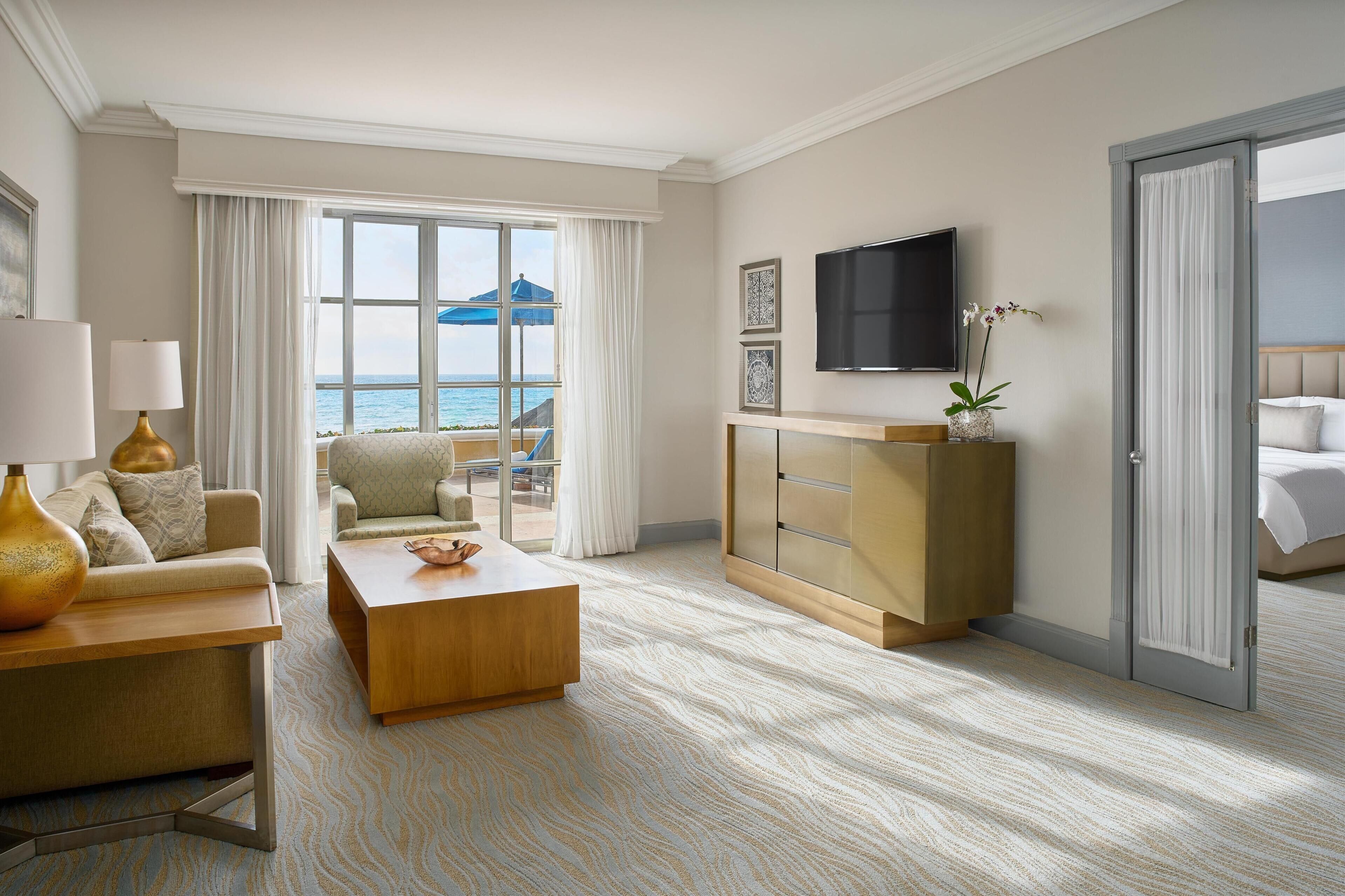 A hotel room with a view of the sea, a sofa, a coffee table, a TV, and a bed.