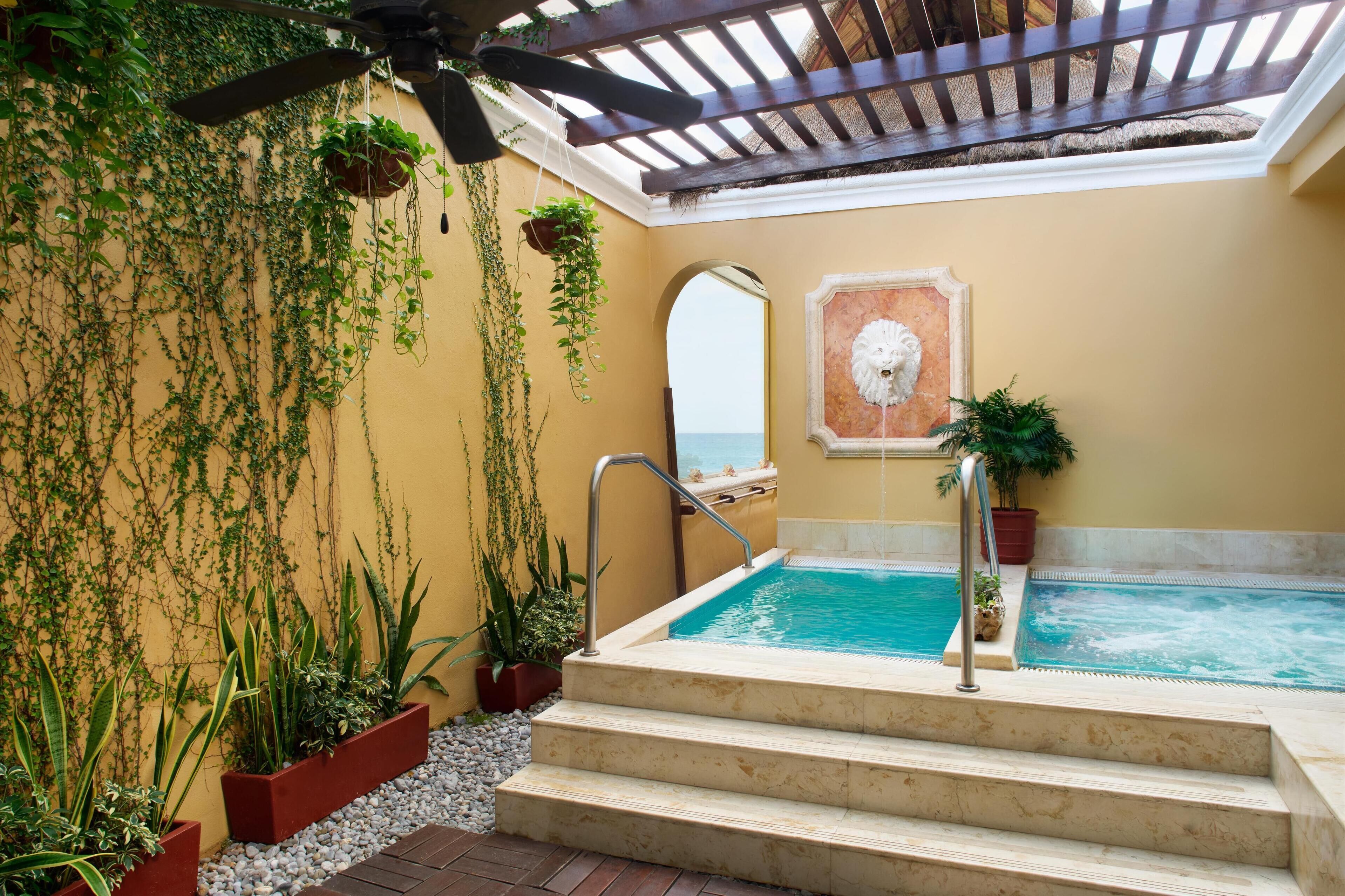A rooftop pool area with steps leading down to a hot tub, surrounded by potted plants and a view of the sea.