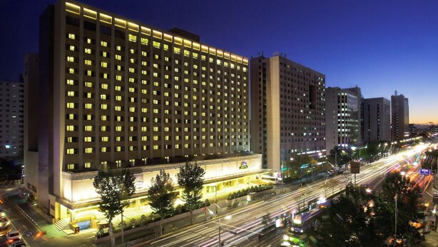 Un hotel de varios pisos con numerosas ventanas, una entrada bien iluminada y una calle concurrida con semáforos y vehículos al frente.