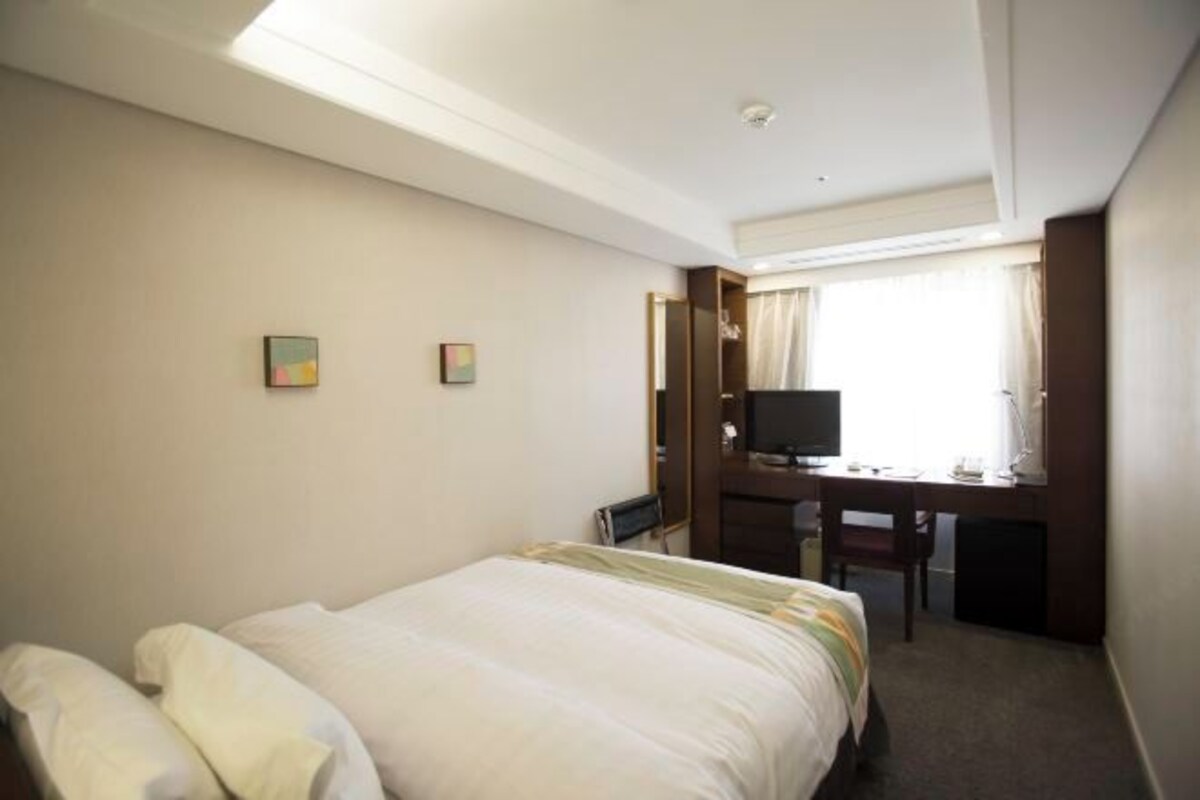 A hotel room with a bed, a desk with a chair, a television, and a window with curtains.