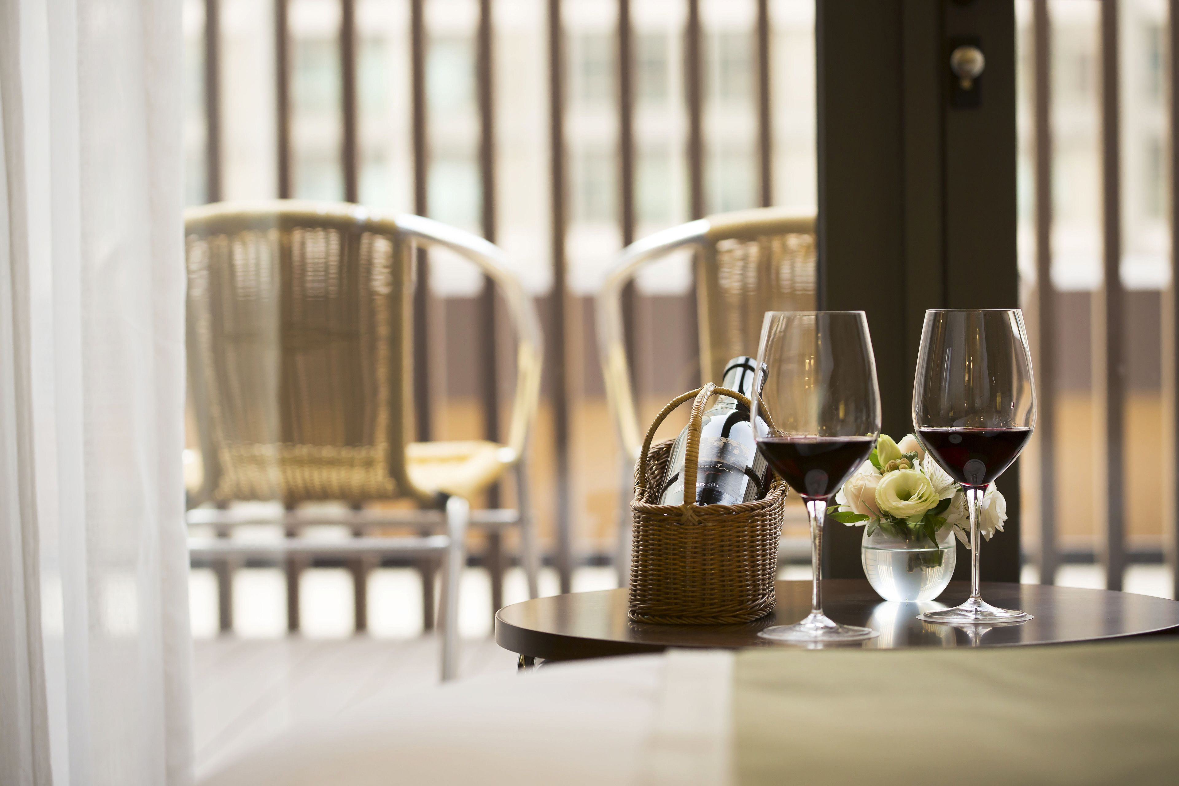 A table with two wine glasses and a wicker basket containing bottles, set on a balcony with wicker chairs and a view of buildings.
