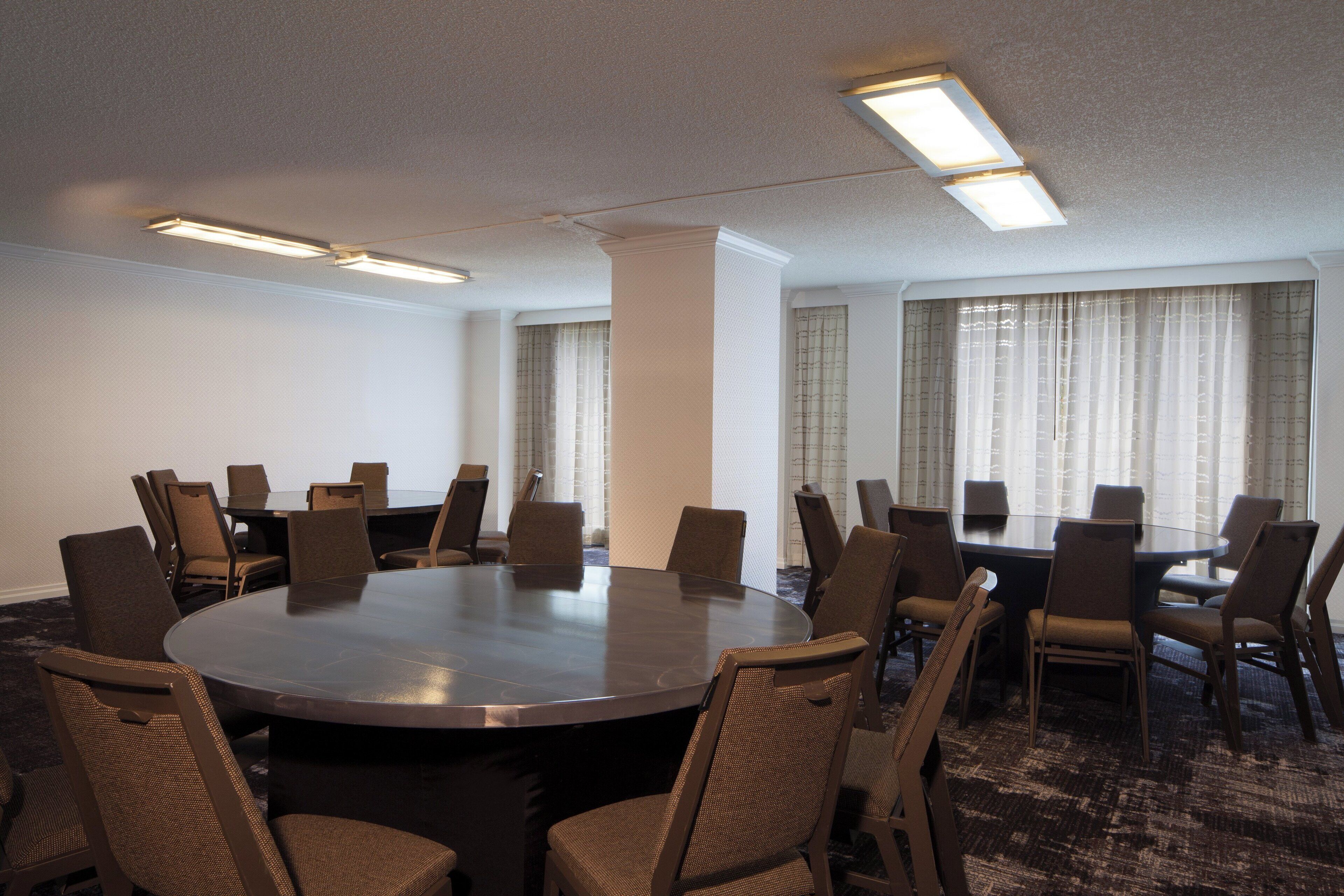 A conference room with a large round table surrounded by chairs, fluorescent ceiling lights, and large windows with curtains.