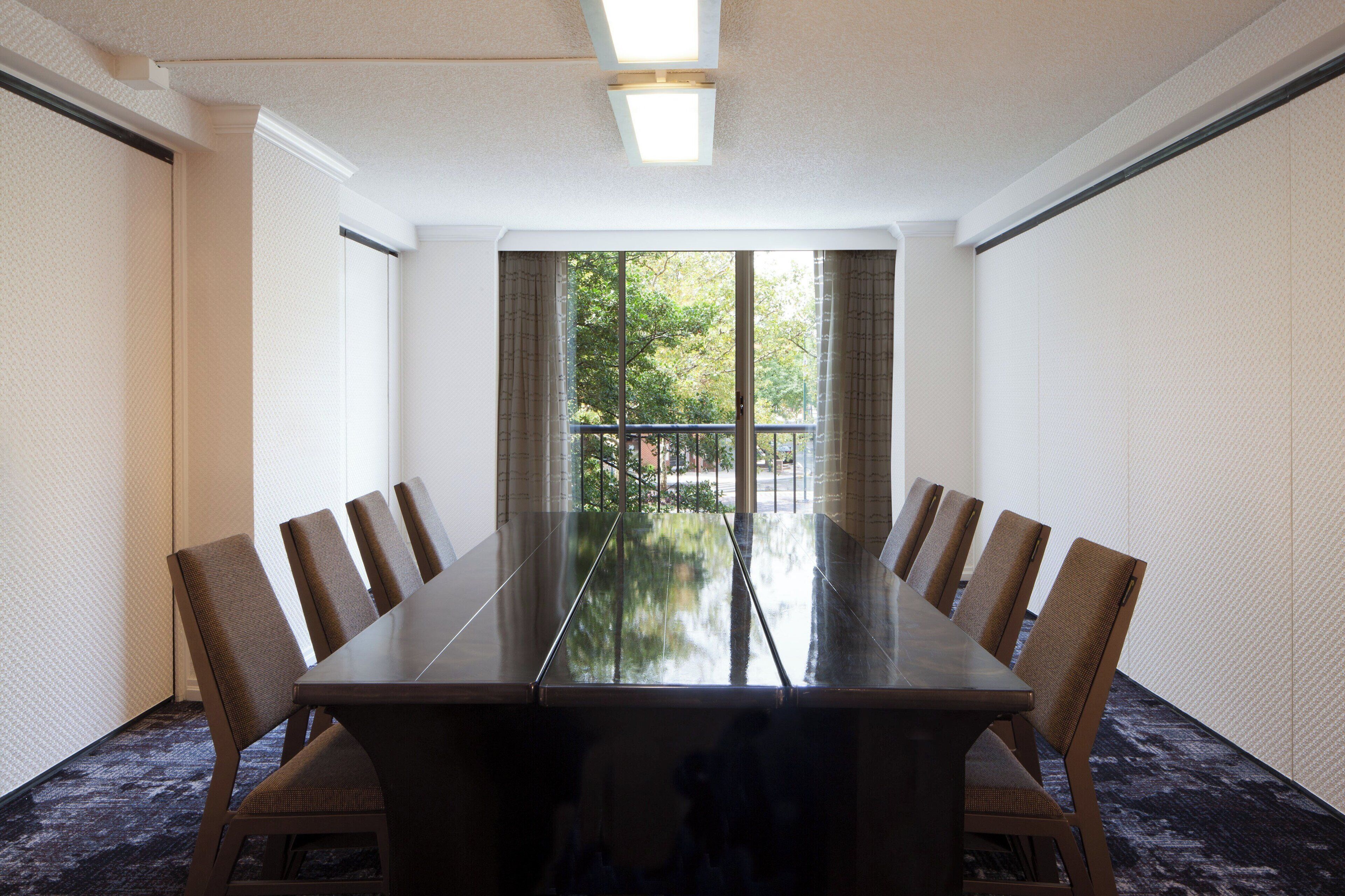 A conference room with a long table and chairs, large windows with curtains, and a carpeted floor.