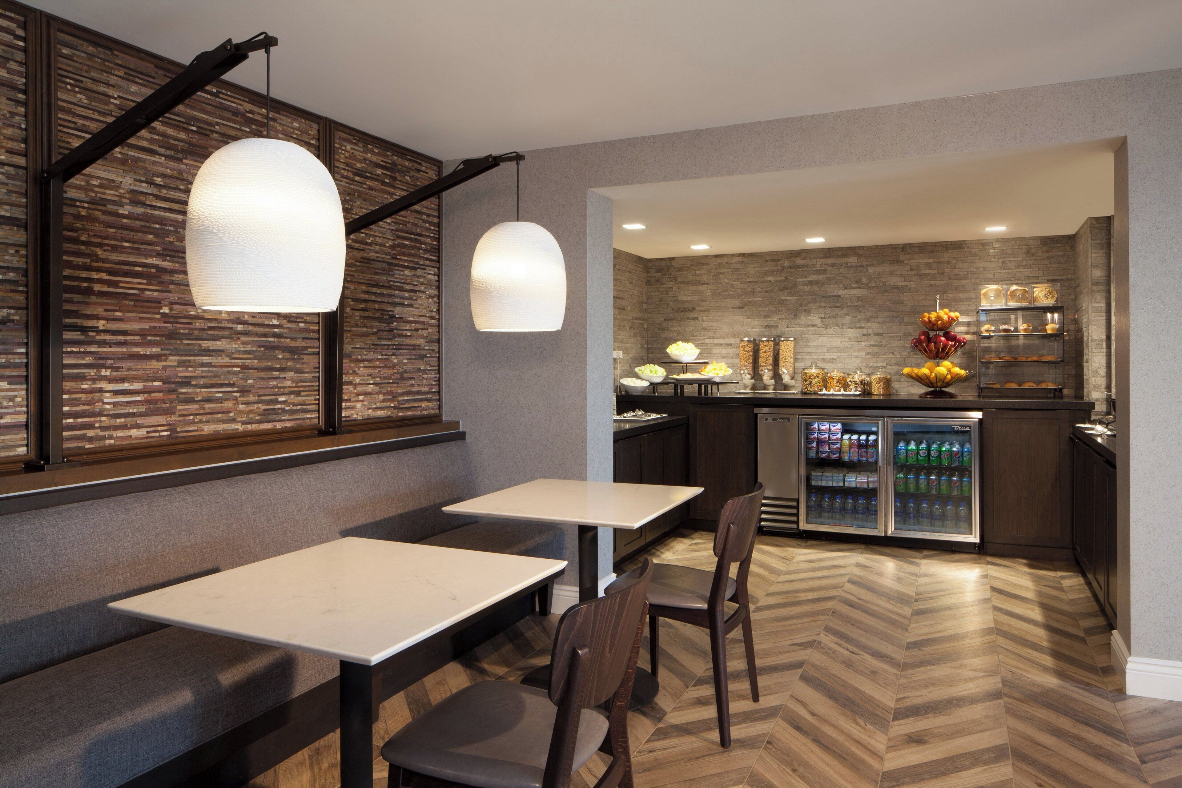 A modern restaurant with a herringbone wood floor, white tables, and dark wooden chairs. A kitchen area with a stone backsplash and a refrigerator stocked with beverages is visible in the background.