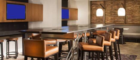 A modern bar area with a long counter, multiple stools, and two flat-screen TVs mounted on the wall.