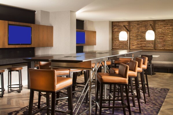 A modern bar area with a long counter, multiple stools, and two flat-screen TVs mounted on the wall.
