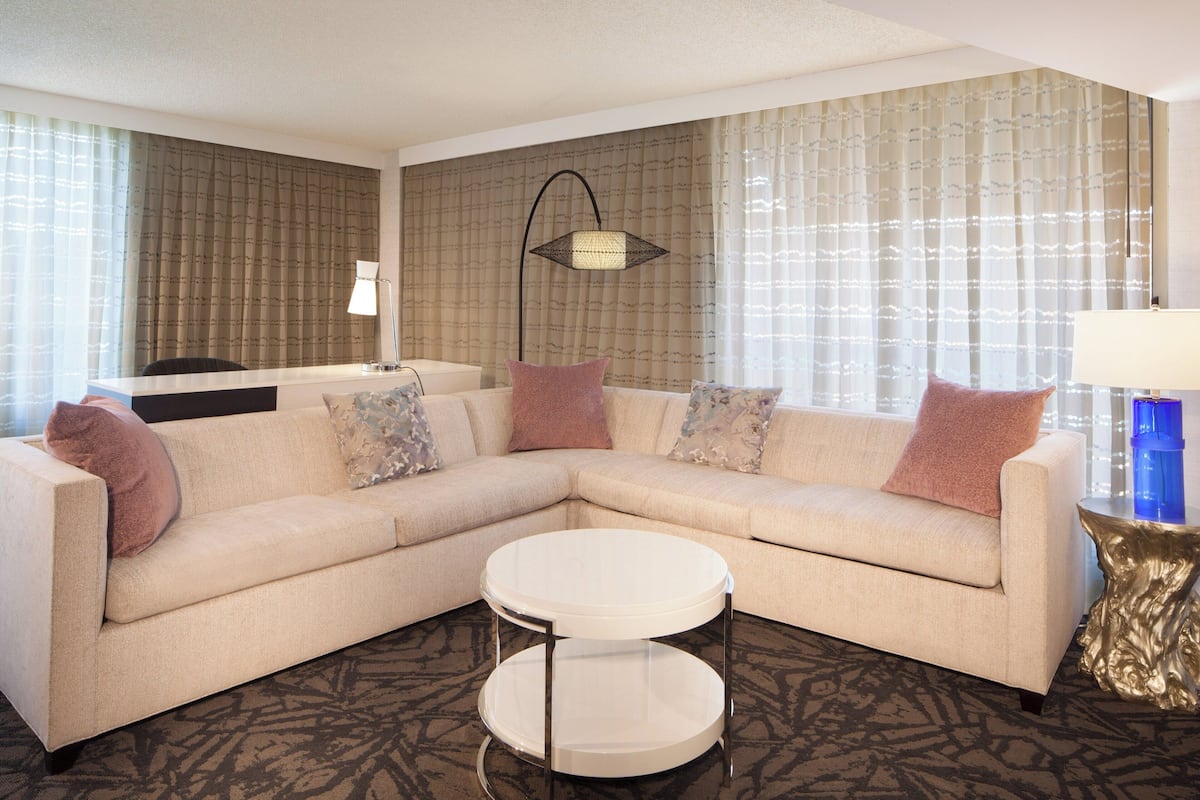 A modern hotel room with a beige sectional sofa, a round coffee table, and a side table with a lamp.