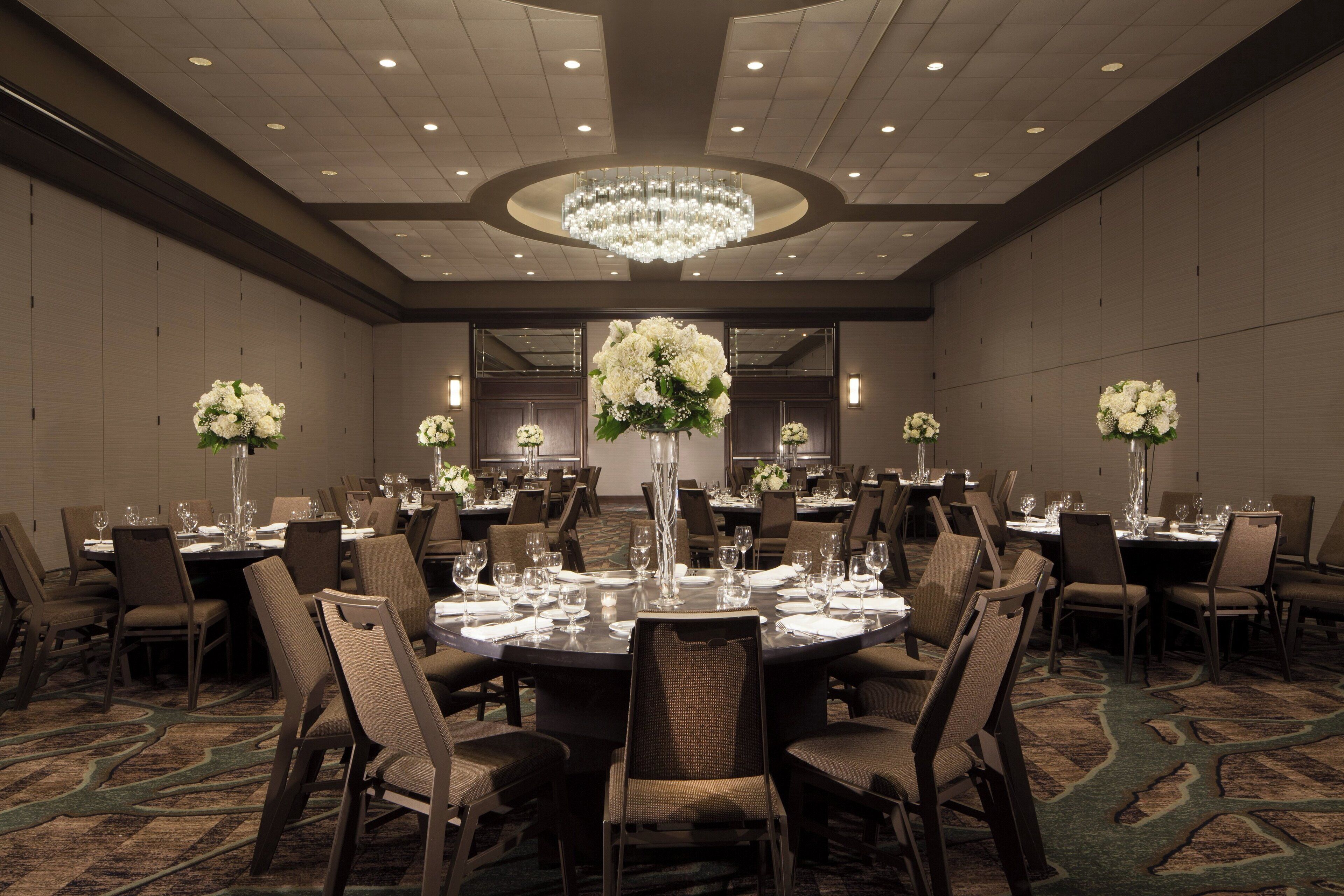 A large, well-lit banquet hall with round tables set for dining, surrounded by chairs, and adorned with tall floral arrangements.