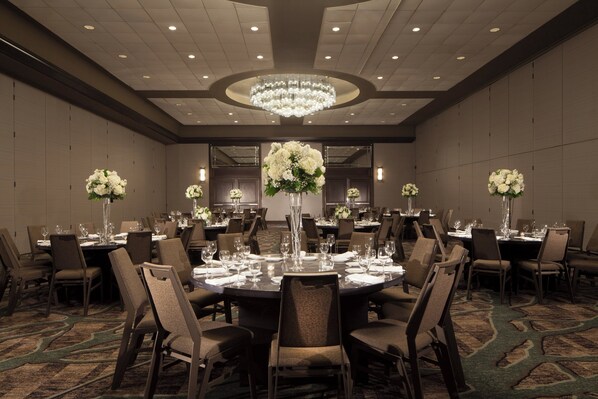 Un amplio salón de banquetes bien iluminado con mesas redondas dispuestas para comer, rodeado de sillas y decorado con altos arreglos florales.