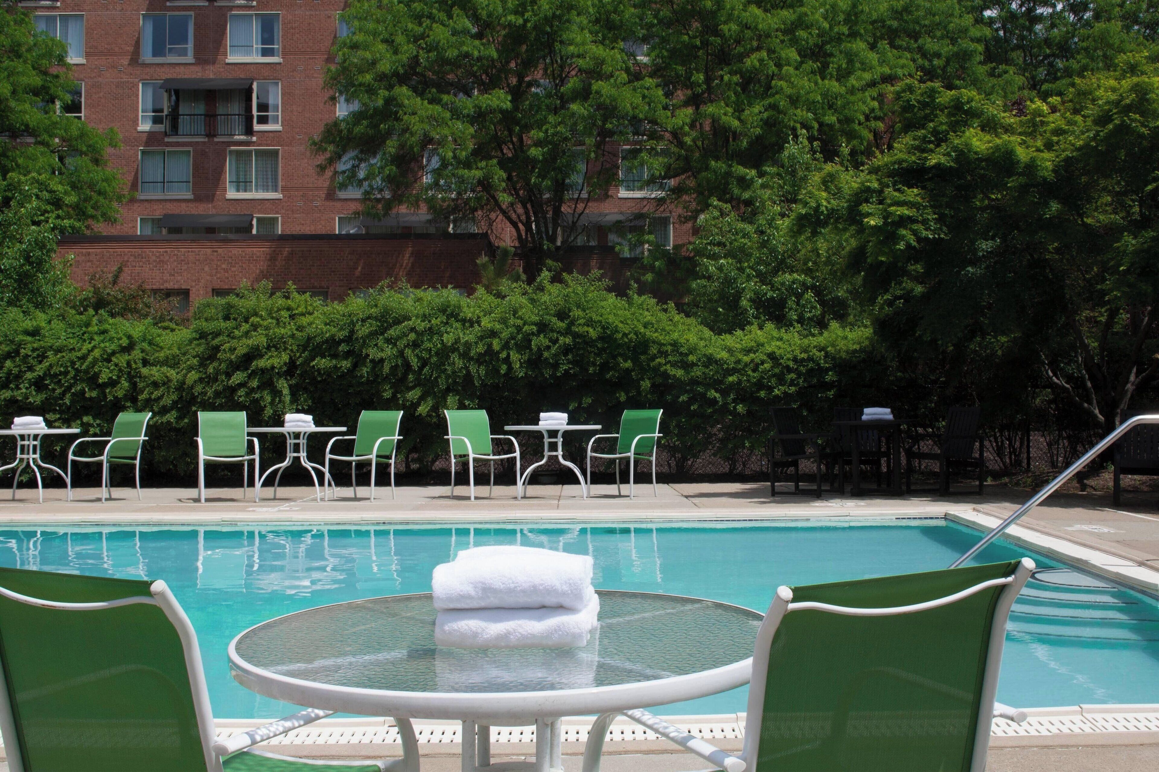 A swimming pool with green chairs and a table, surrounded by trees and a brick building in the background.