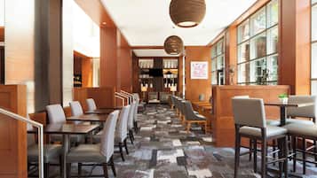 A hotel lobby with a long reception desk, multiple seating areas with chairs and tables, a patterned carpet, wooden walls, and large windows.