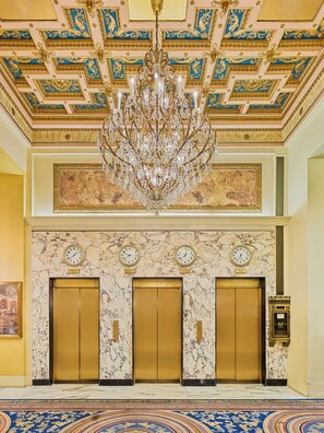 A grand lobby with a large chandelier, marble walls, and three elevators.