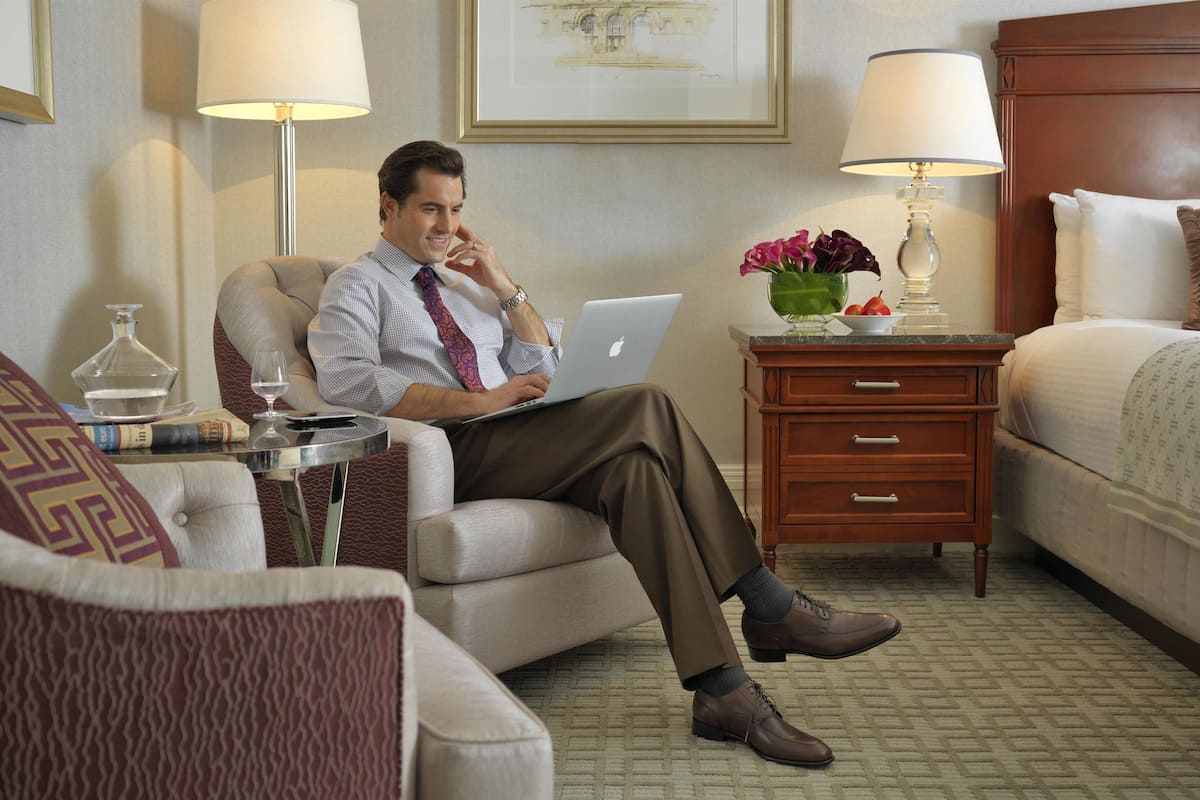 A hotel room with a bed, a sitting area with a chair, a desk with a laptop, a nightstand with a lamp, and a framed picture on the wall.