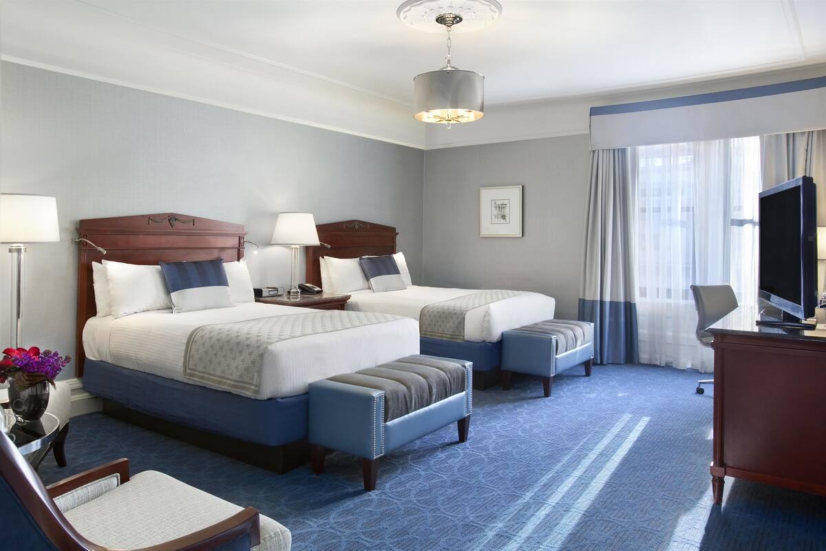 A hotel room with two beds, a desk, a chair, and a television.
