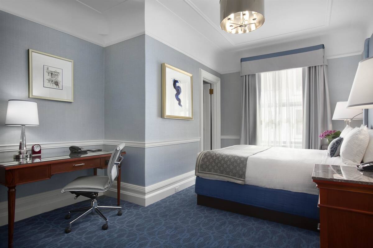 A hotel room with a bed, desk, chair, and a large window with curtains.
