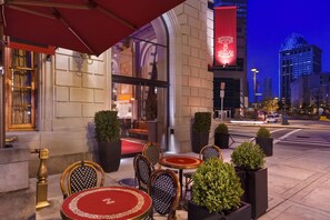 An outdoor seating area with tables and chairs, a red awning, and a building entrance with a red banner.