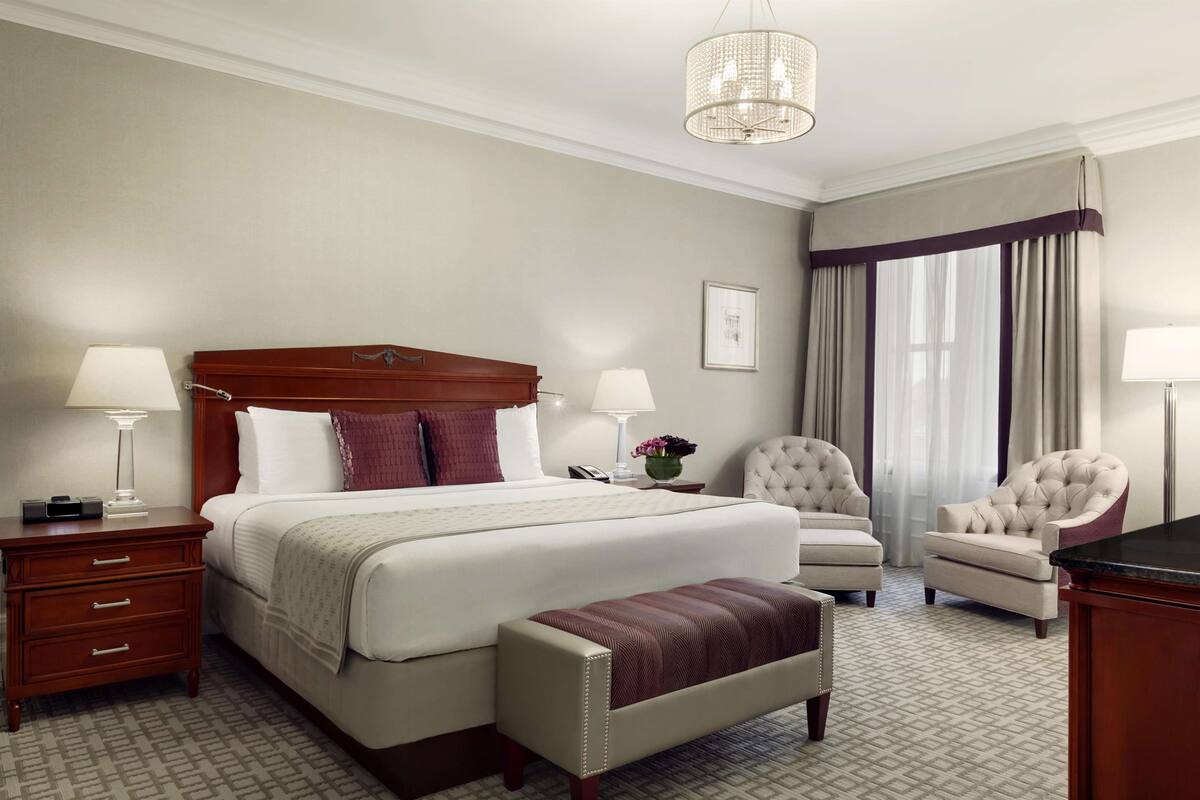A hotel room with a large bed, two bedside lamps, a wooden nightstand, a bench, a chair, and a desk.