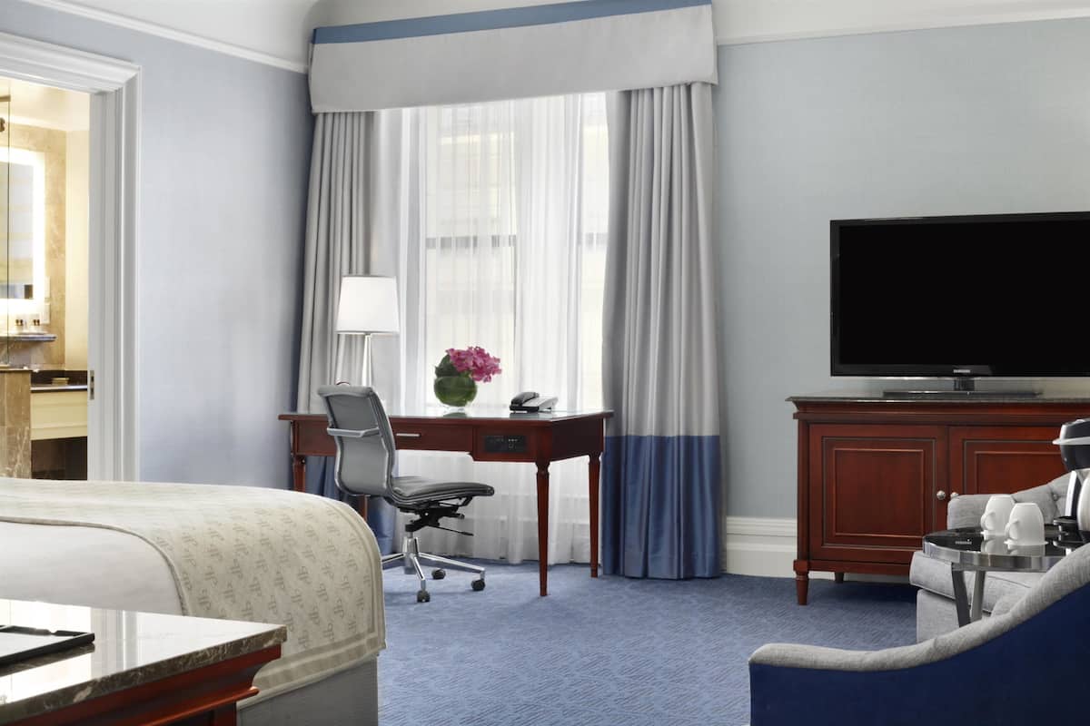 A hotel room with a bed, a desk with a chair, a television, and a blue armchair.