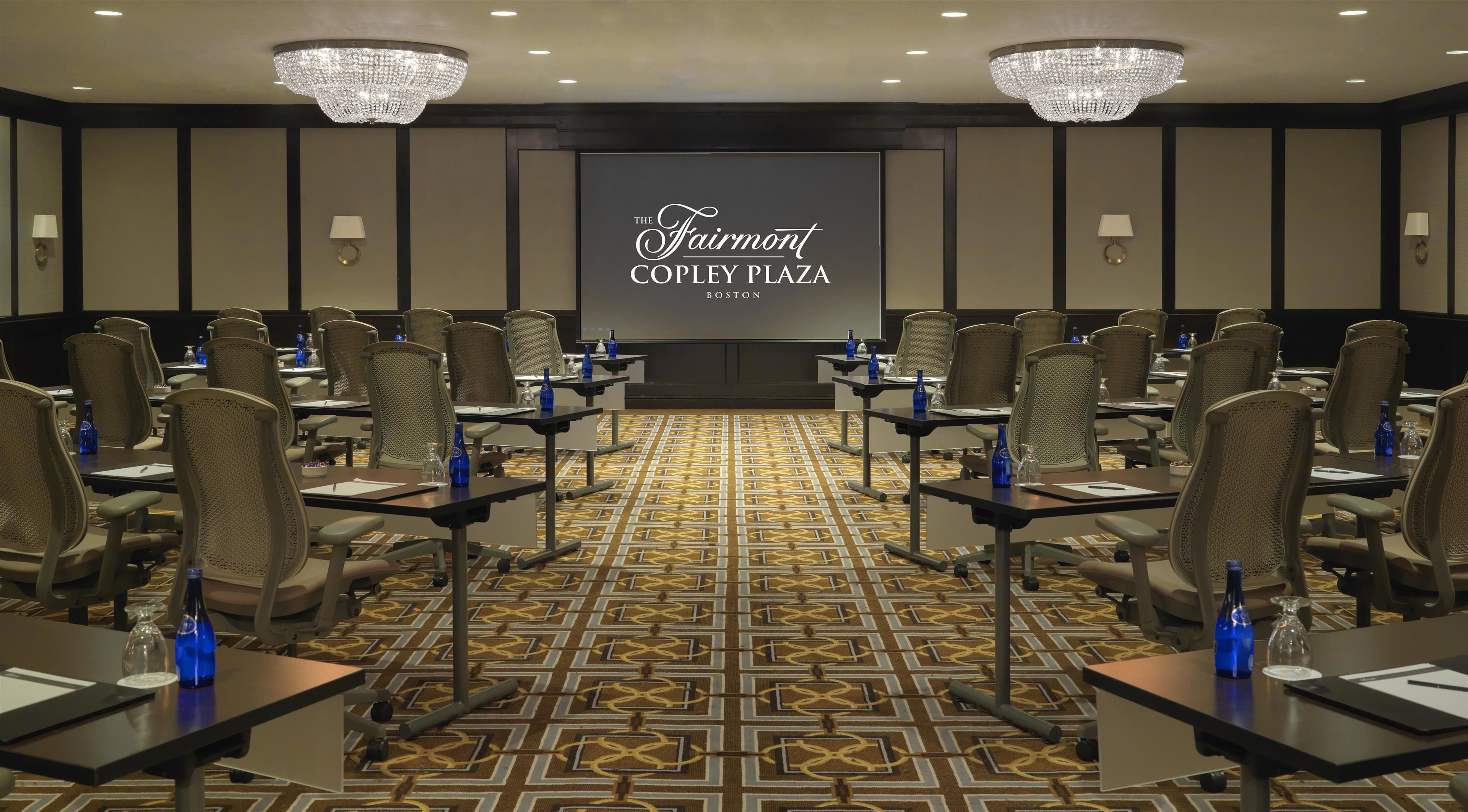 Un salón de conferencias en The Fairmont Copley Plaza, Boston, con filas de sillas, mesas y una pantalla grande que muestra el nombre del hotel.