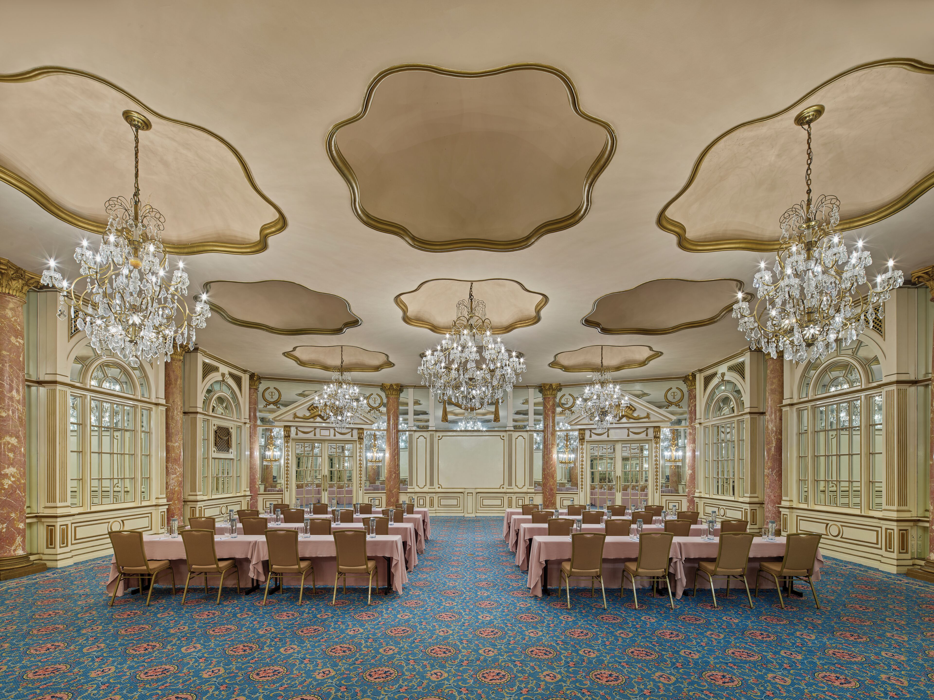 A grand ballroom with ornate chandeliers, large tables, and a patterned carpet.