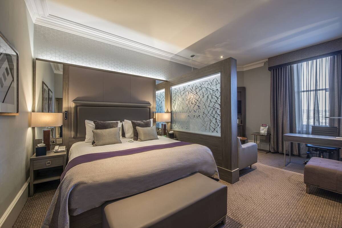 A hotel room with a large bed, a desk, a chair, and a window with curtains.