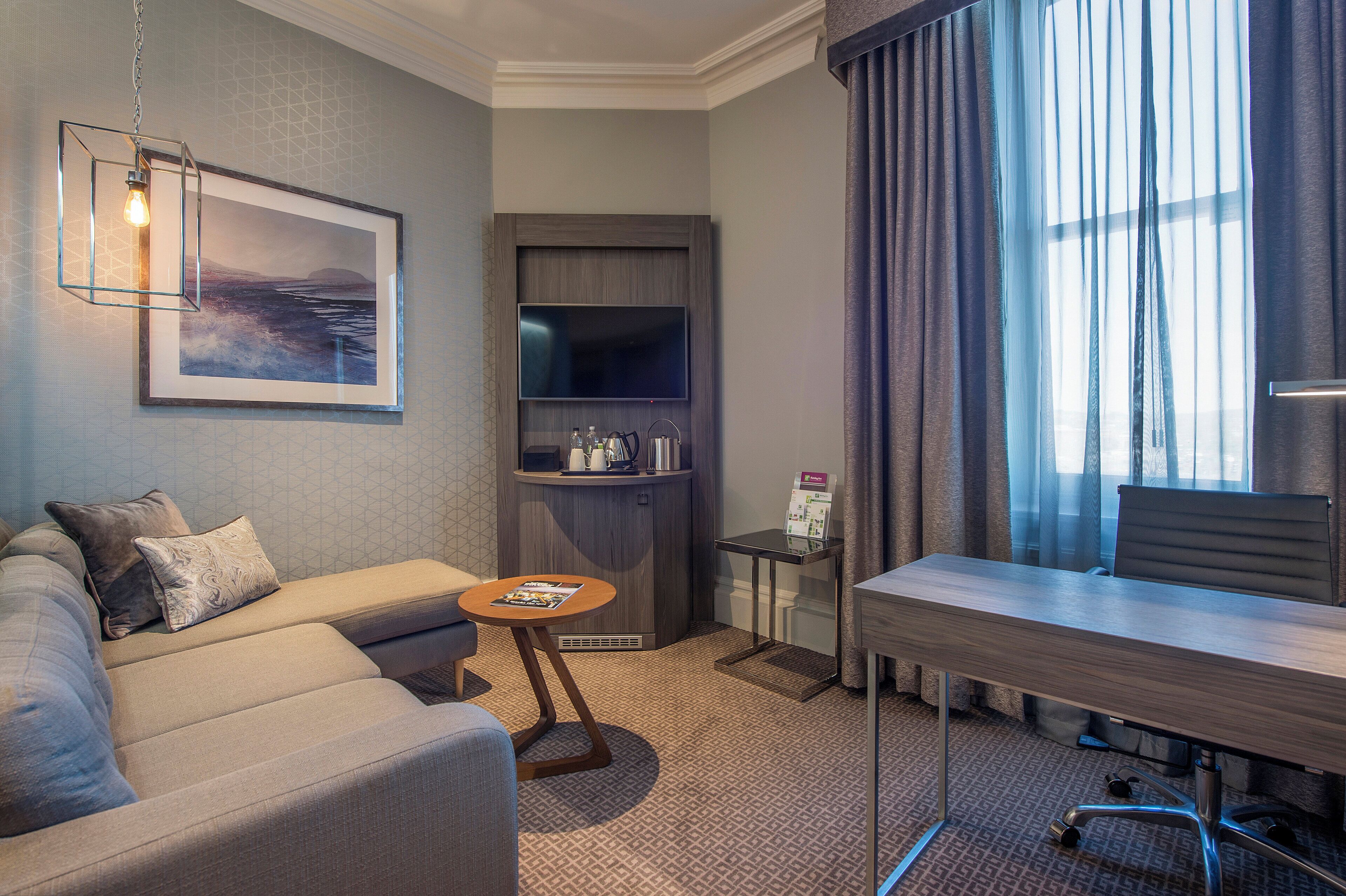 Ein Hotelzimmer mit Sofa, einem kleinen Tisch, einem Schreibtisch, einem Fernseher und einem Fenster mit Vorhängen.