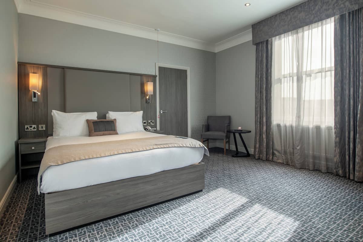 A modern hotel room with a large bed, a nightstand with a lamp, a chair, and a view of a cityscape through sheer curtains.