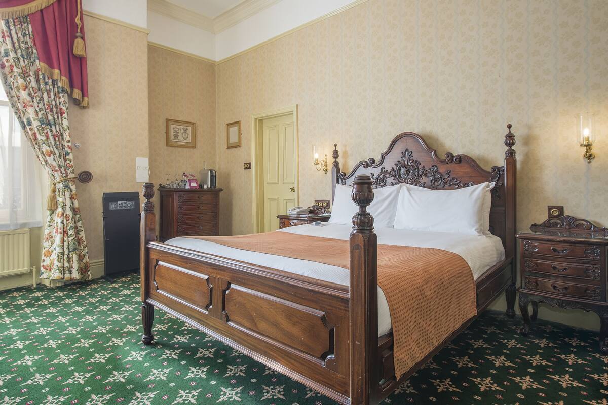 A classic four-poster bed with a wooden frame and a green patterned carpet. There is a nightstand with a lamp and a dresser with a mirror.