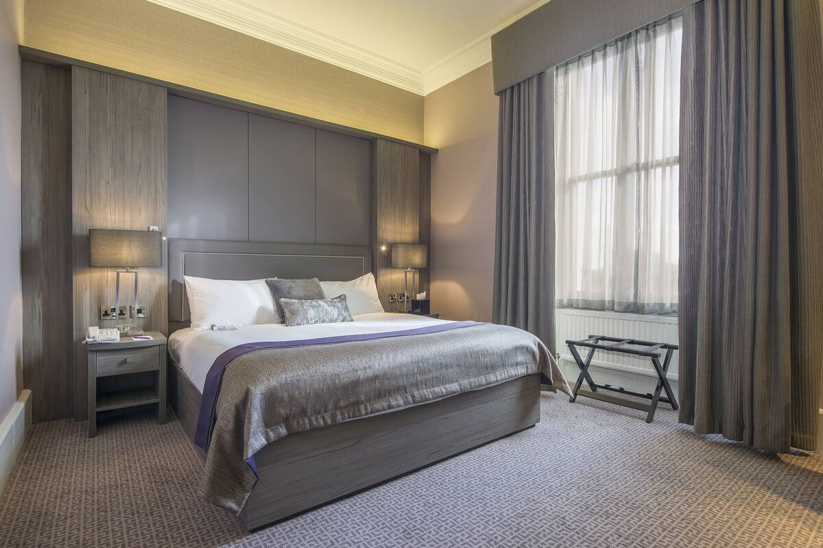 A modern hotel room with a large bed, bedside tables, a nightstand, a window with curtains, and a radiator.
