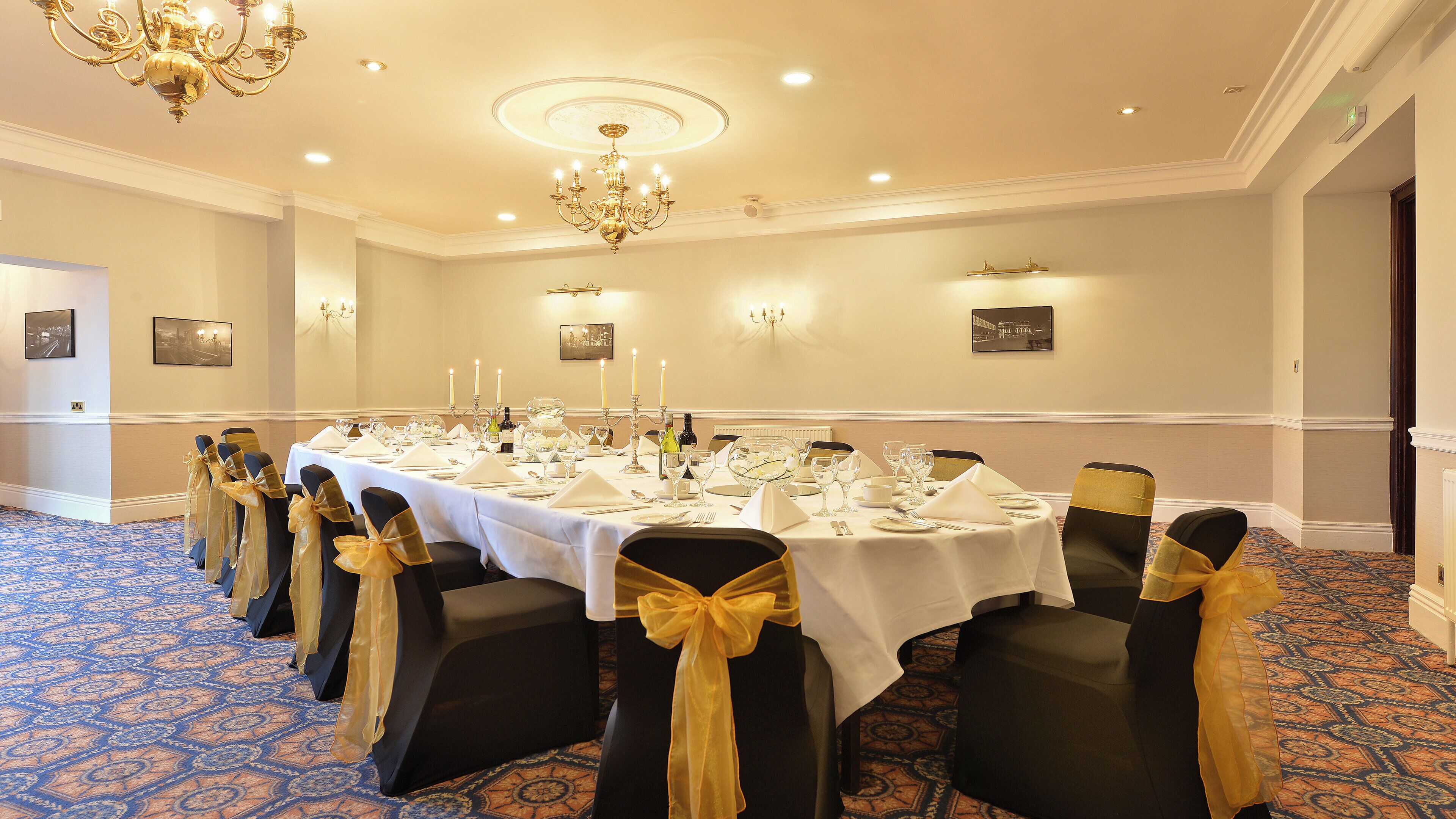 A banquet hall with a long table set for a formal event, featuring chairs with gold sashes, a chandelier, and wall-mounted lighting.