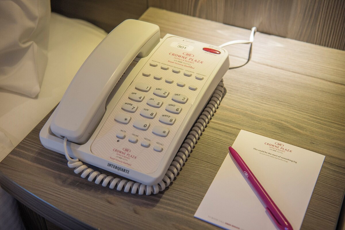 Un téléphone blanc, posé sur une table de chevet en bois, avec un bloc-notes et un stylo à côté.