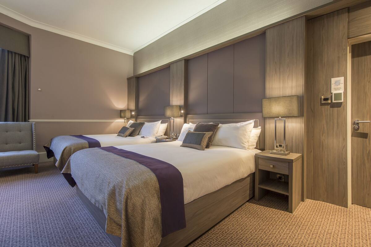 A hotel room with two beds, a chair, and a nightstand with a lamp.