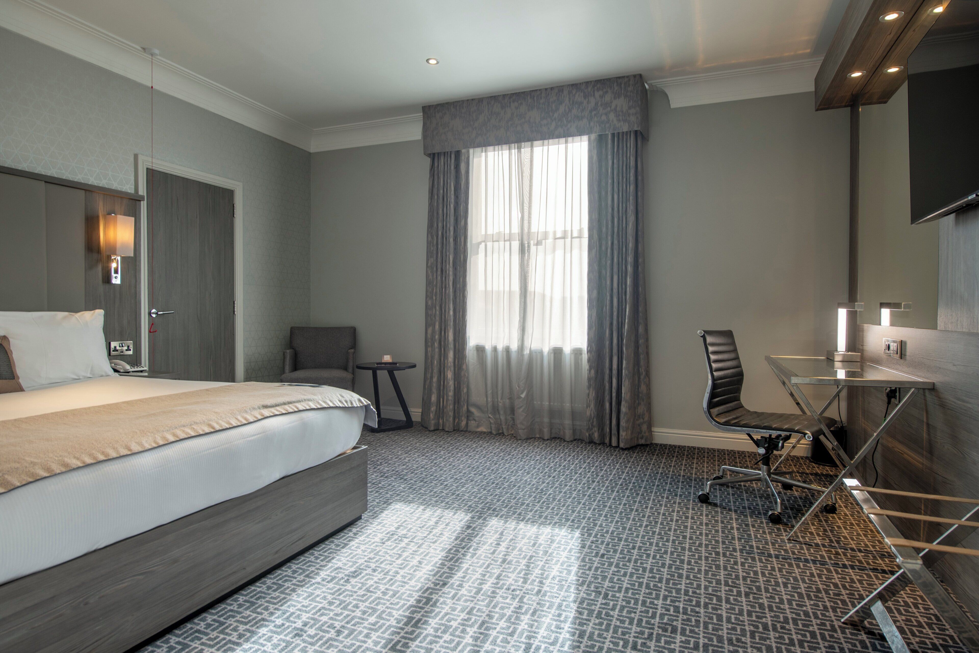 A hotel room with a large bed, a desk with a chair, a small table, and a large window with curtains.