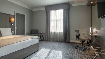 A hotel room with a large bed, a desk with a chair, a small table, and a large window with curtains.