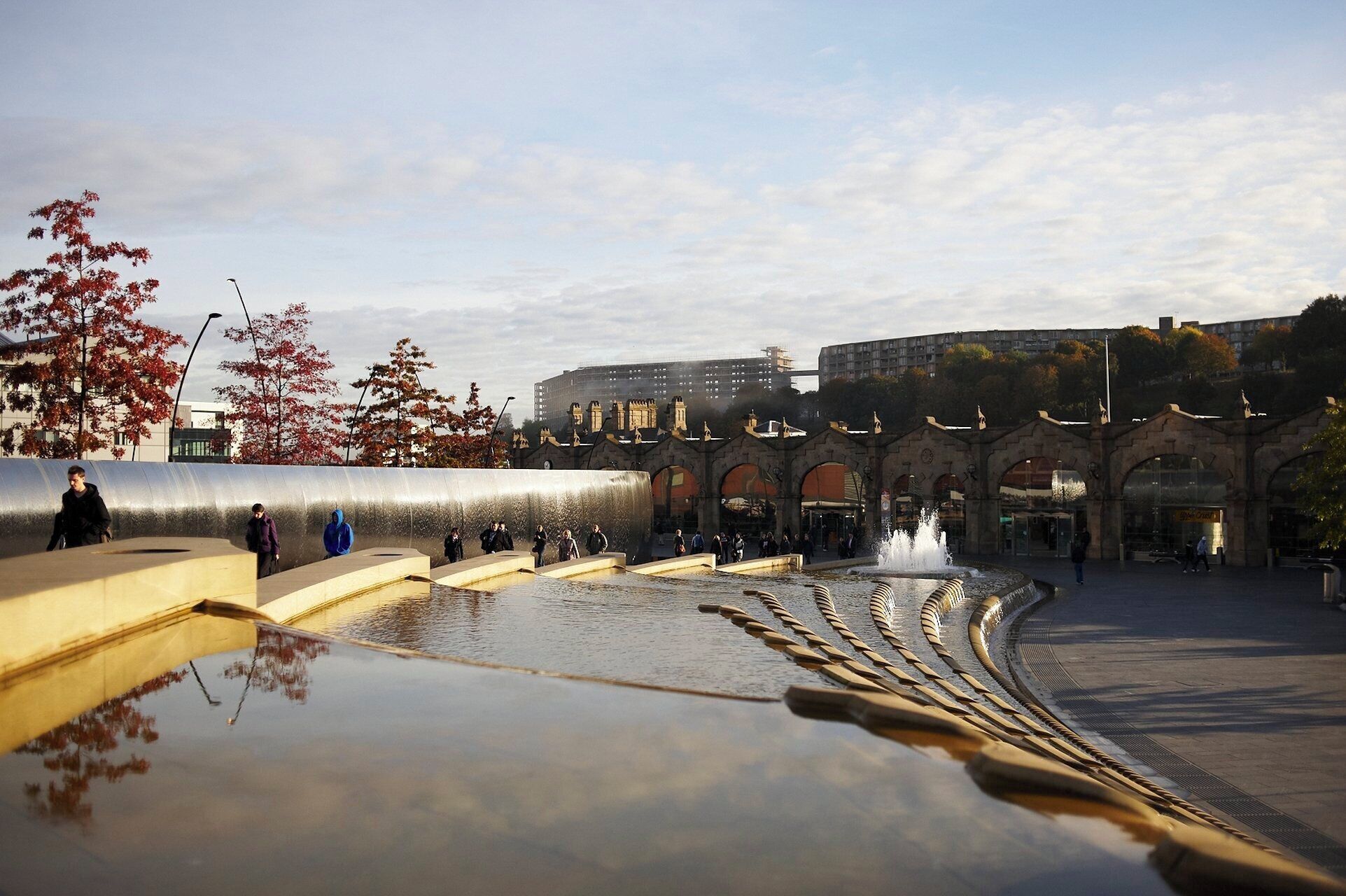 A public square with a reflective water feature and a historic building with arches.