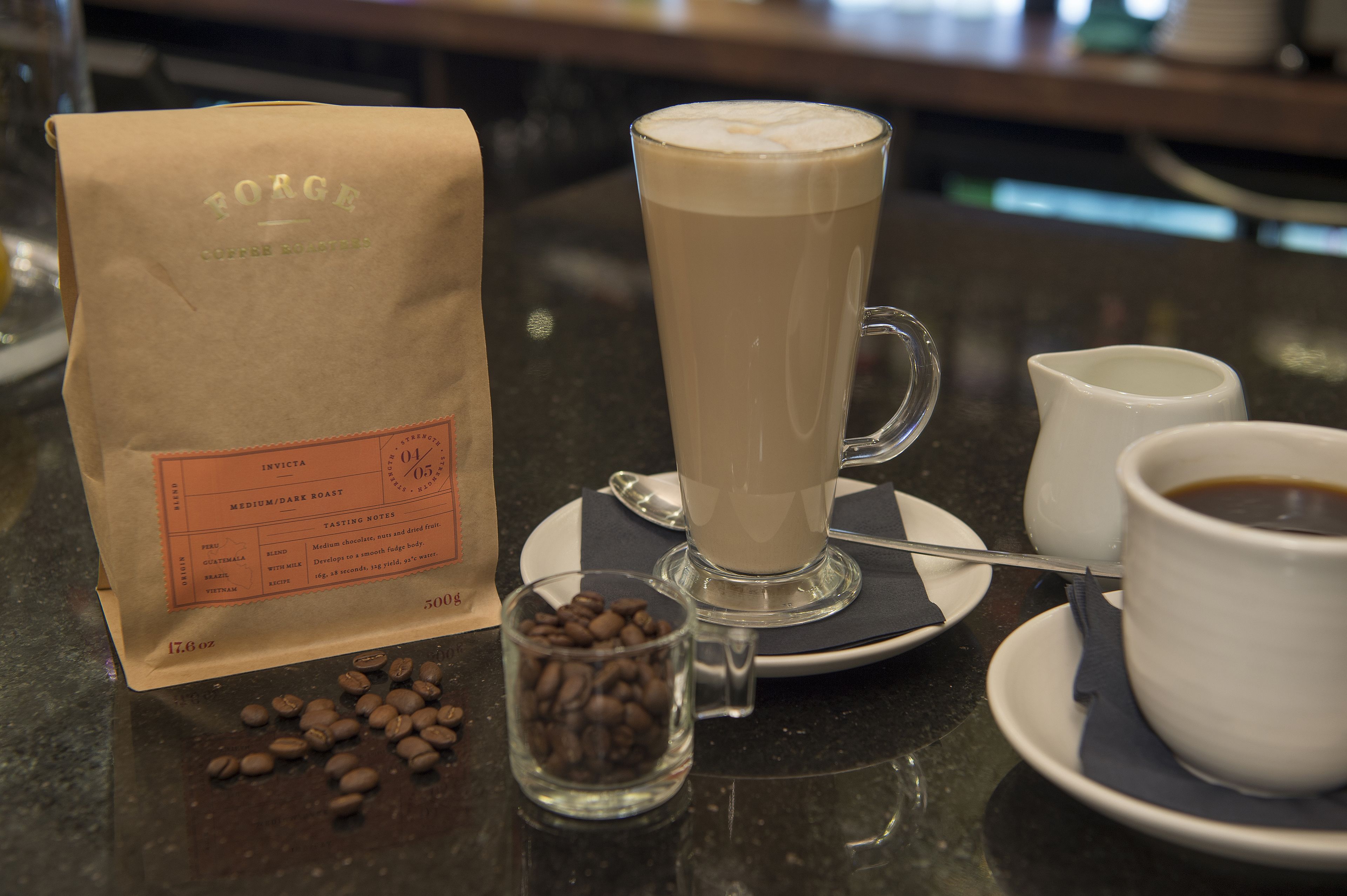 Una bolsa de café, una taza alta de café, un vaso pequeño con granos de café y una taza de café sobre una mesa.