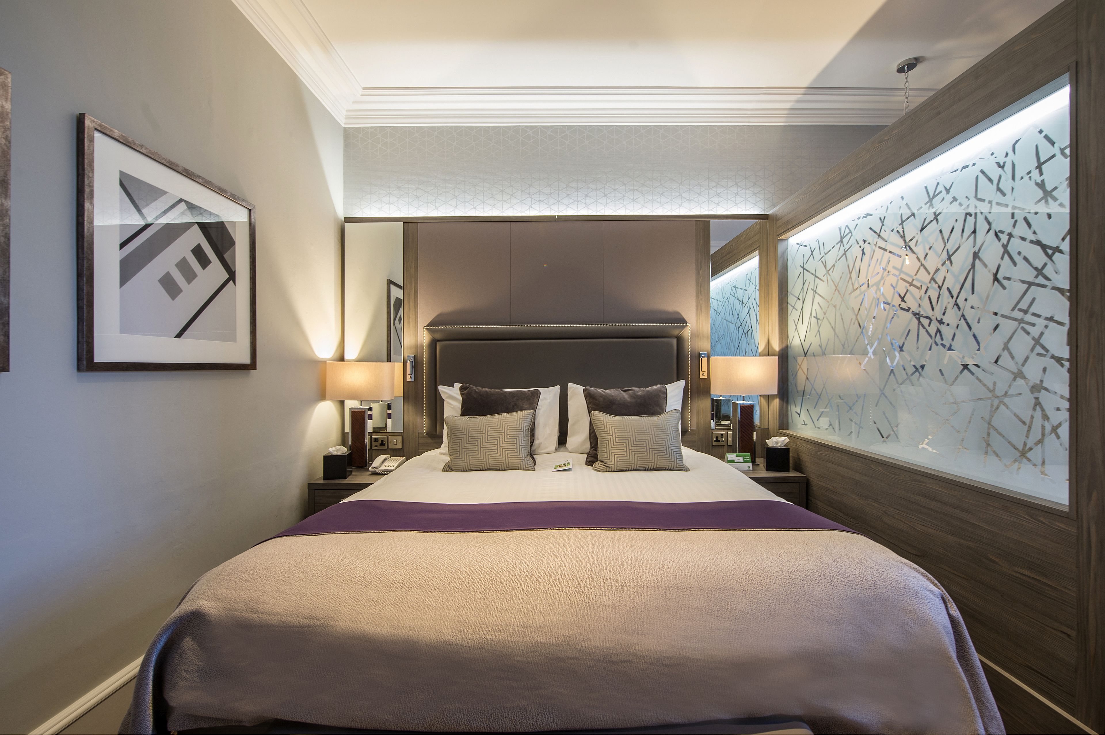 A hotel room with a large bed, two bedside tables with lamps, a framed artwork on the wall, and a decorative wall panel.