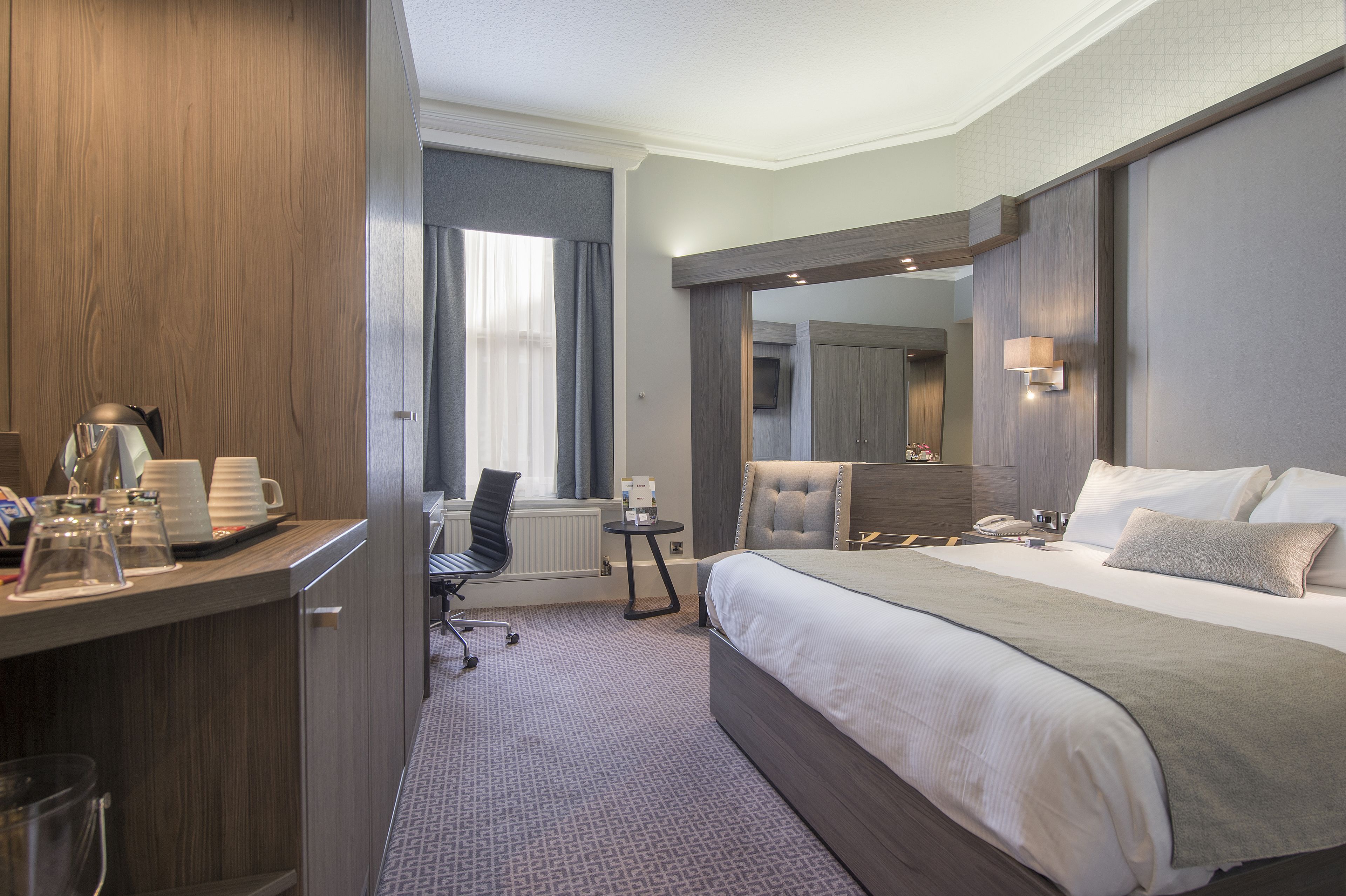 Ein Hotelzimmer mit einem großen Bett, einem Schreibtisch mit Kaffeemaschine, einem Sessel, einem kleinen Tisch und einem Spiegel.