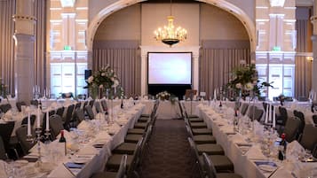 A banquet hall with long tables set for a formal event, featuring chandeliers, floral centerpieces, and a large screen at the front.
