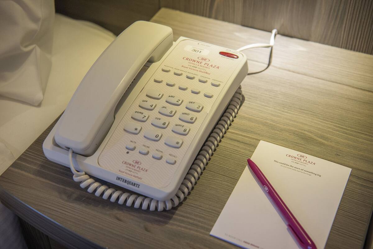 A white hotel phone on a wooden bedside table with a notepad and pen beside it.