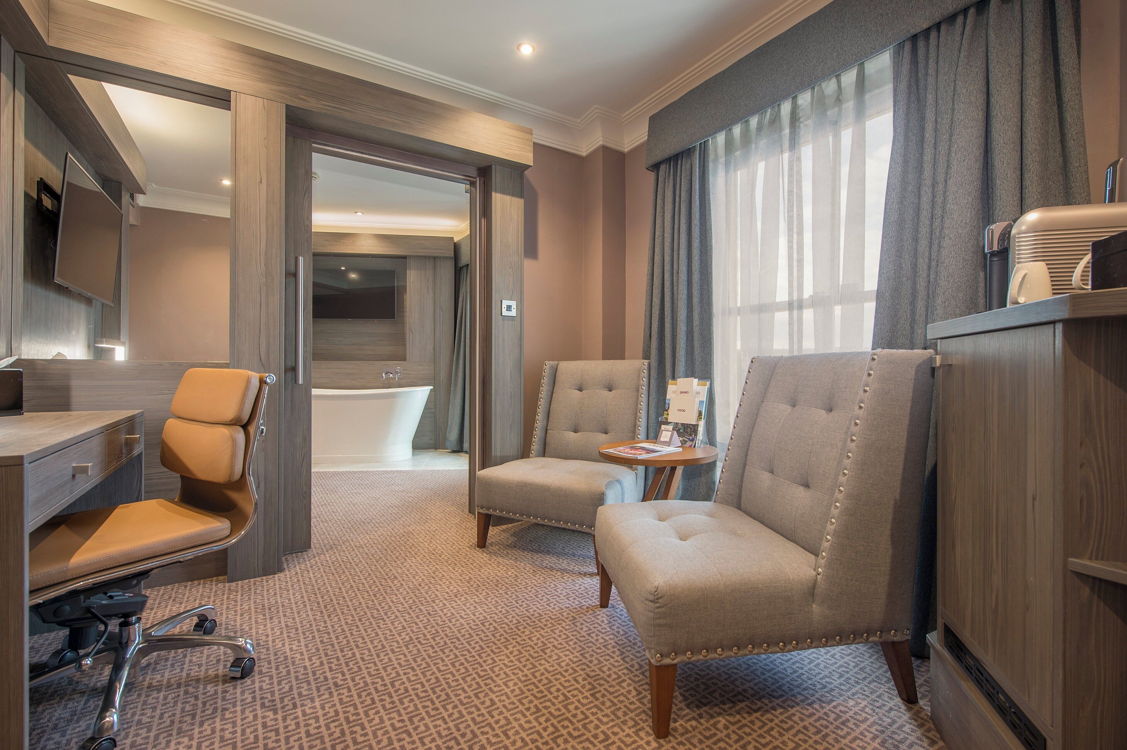 A hotel room with a desk, two armchairs, a small table, a bathtub, and a minibar.