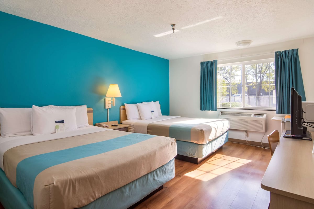 Une chambre d’hôtel avec deux lits, un bureau, une chaise, une lampe et une fenêtre avec des rideaux.