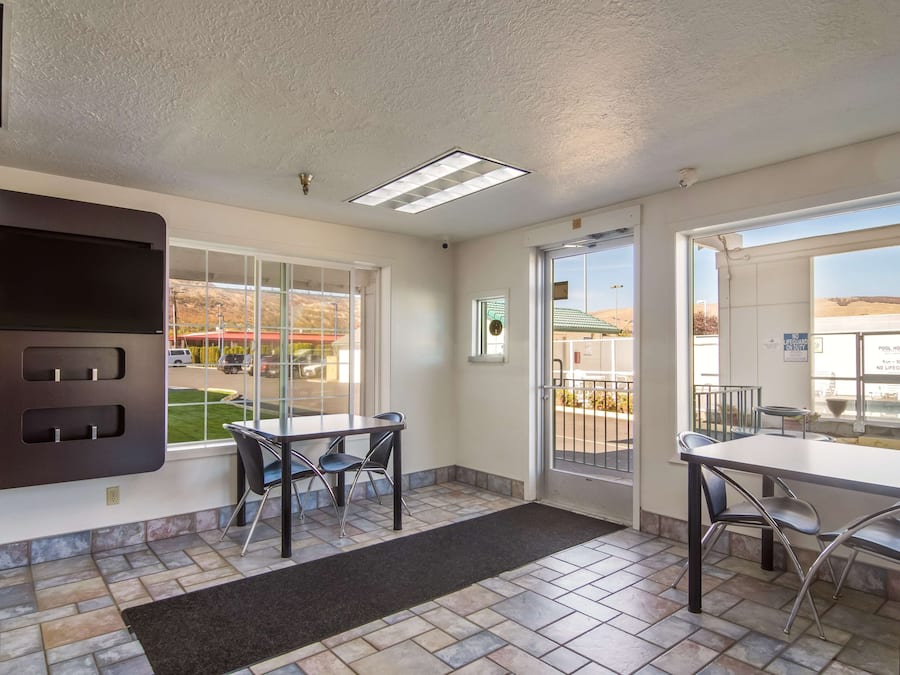 A hotel room with a large window, a flat-screen TV mounted on the wall, a dining area with a table and chairs, and a tiled floor.
