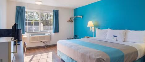 Une chambre d’hôtel avec un grand lit, un bureau avec un ordinateur, une fenêtre avec des rideaux et une lampe fixée au mur.